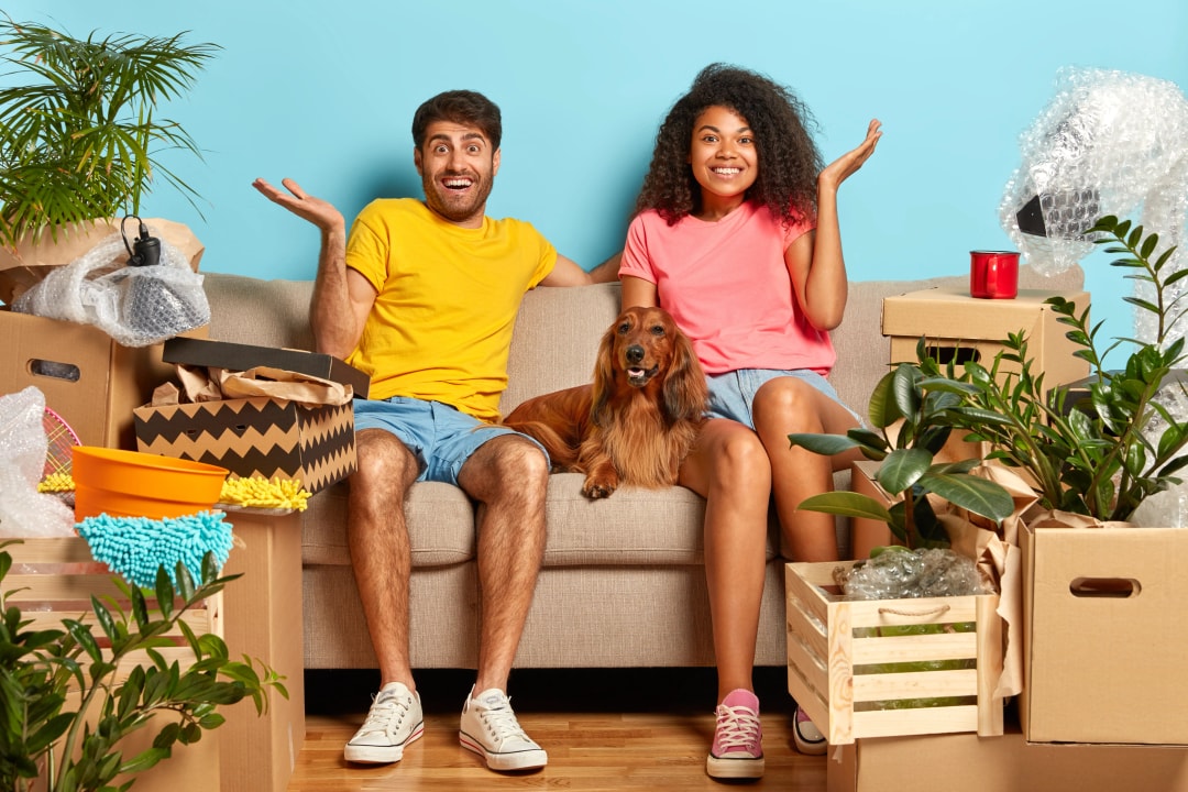 Jeune couple assis sur un canapé avec leur chien, entouré de cartons de déménagement, en tant que primo-accédants