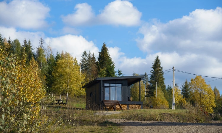 Tiny-house-en-bois-située-en-pleine-nature,-entourée-d'arbres-aux-couleurs-automnales-sous-un-ciel-bleu-avec-quelques-nuages.